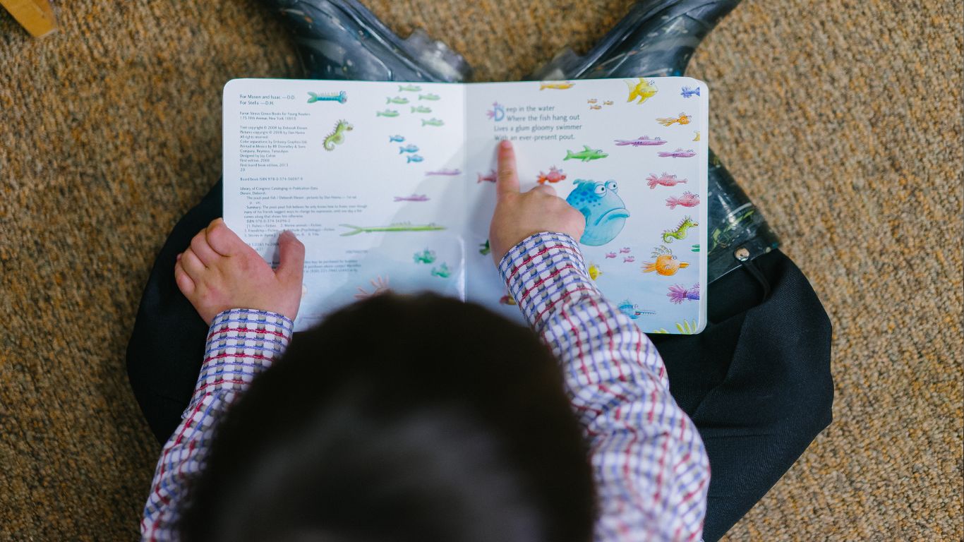 An elementary student points out the words as he reads while sitting on the floor of a school library.